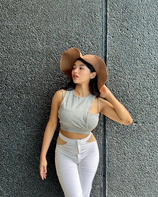 Woman wearing a gray crop top and white pants with a brown hat against a textured gray wall.