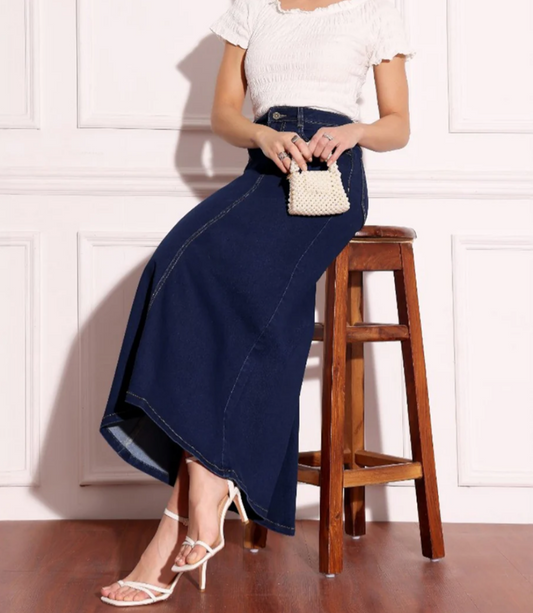 Woman wearing a navy blue denim skirt and white top, sitting on a wooden stool against a white wall.