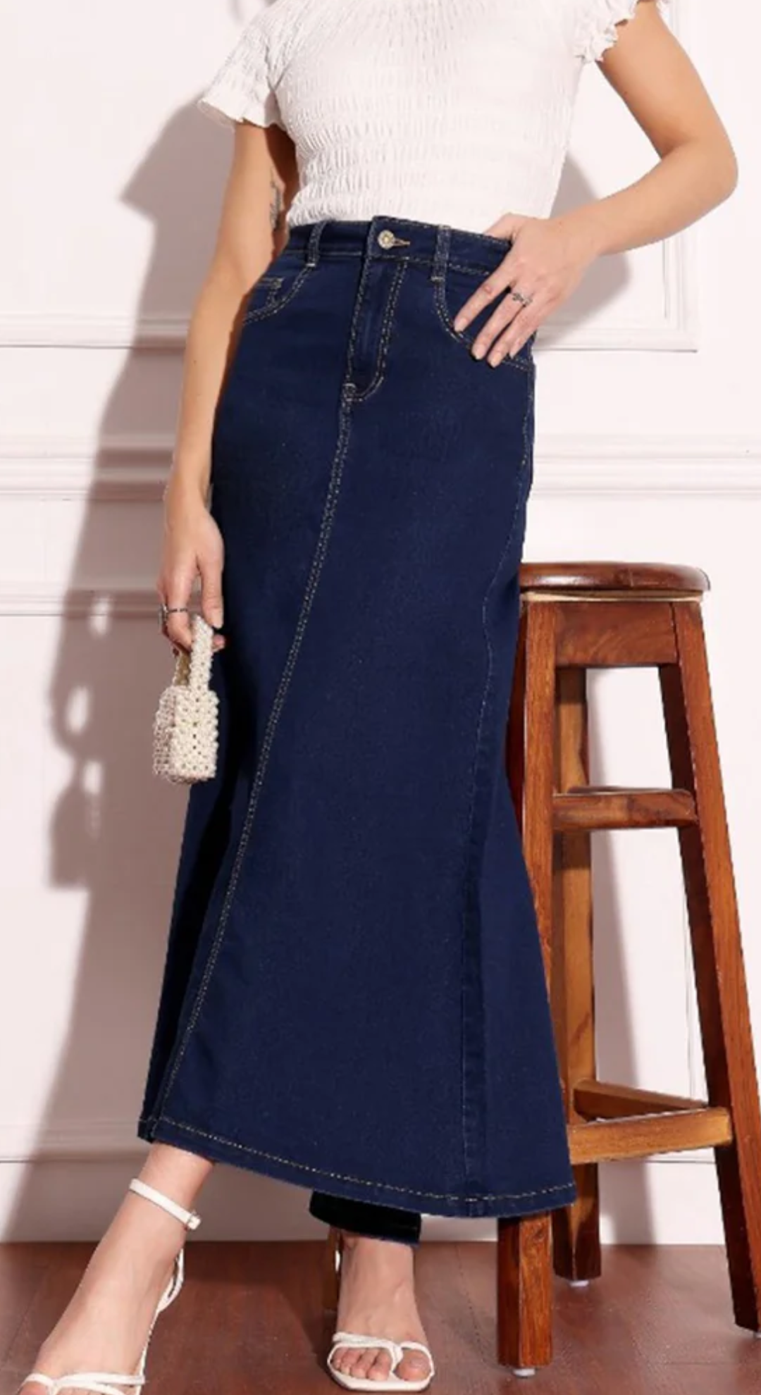 Person wearing a long navy denim skirt with a white top and sandals, standing next to a wooden stool.