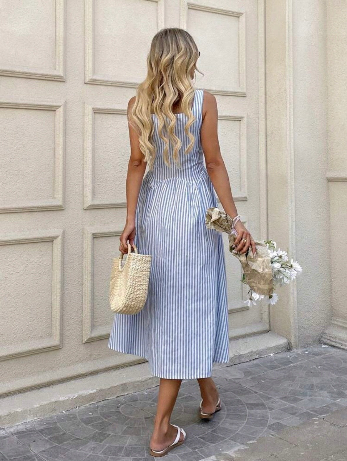 Woman in a blue striped dress holding a straw bag and flowers, standing in front of a building entrance.