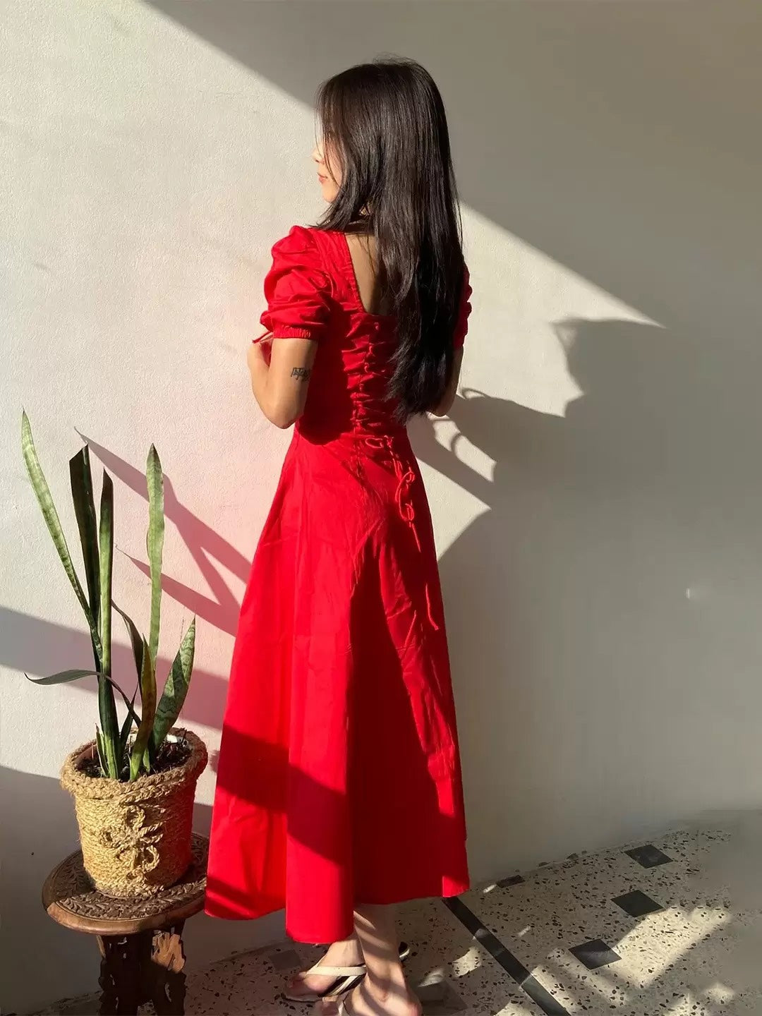 Woman in a red dress standing against a white wall with a plant on a small table beside her.