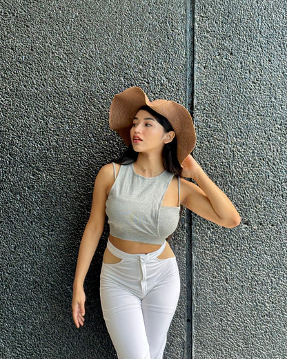 Woman wearing a gray crop top and white pants with a brown hat against a textured gray wall.