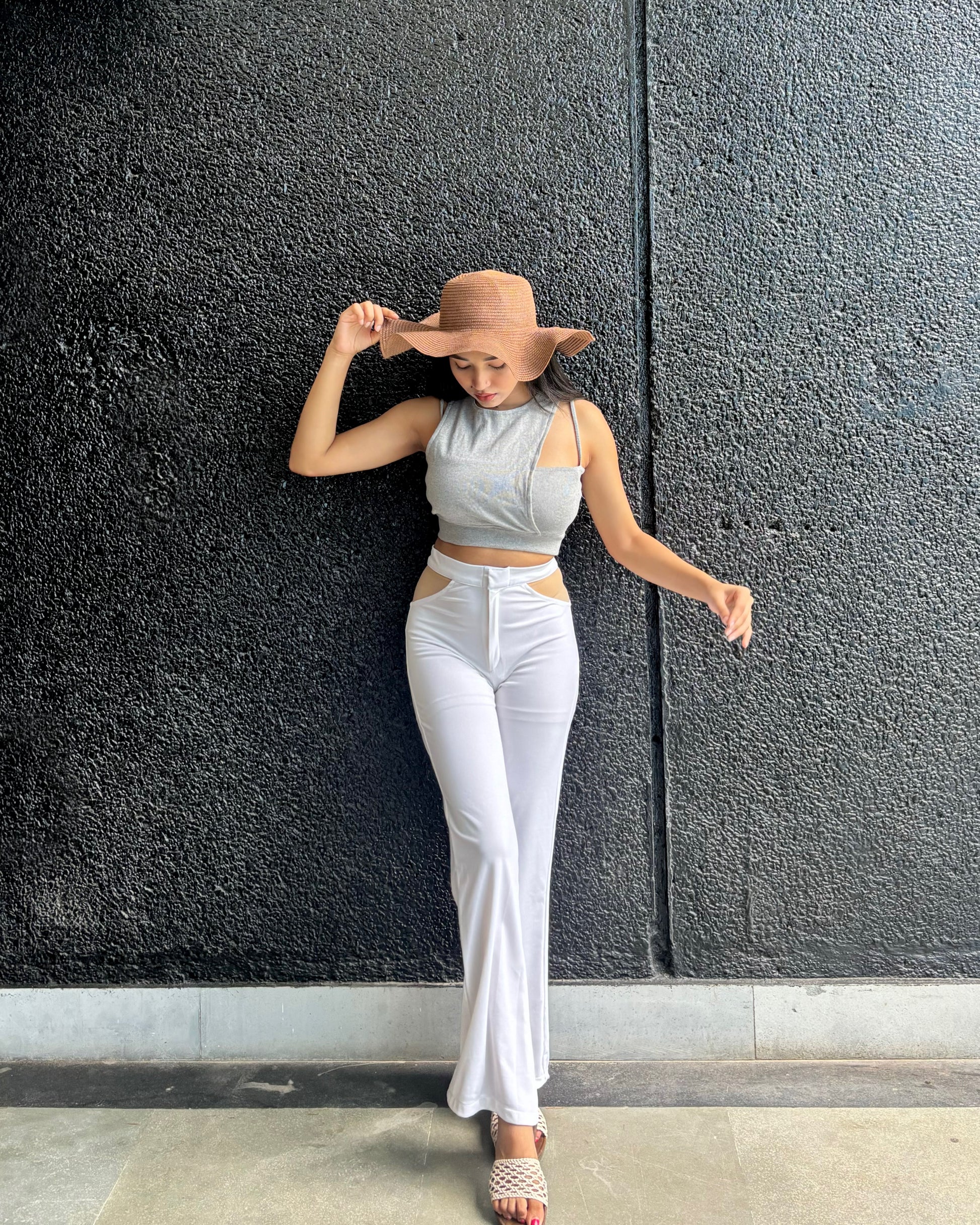 Woman wearing a light gray top and white pants against a dark textured wall.