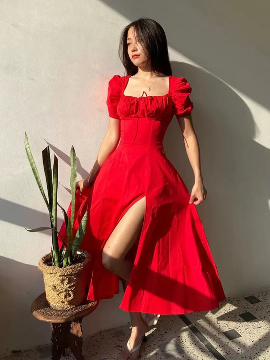 Woman in a red dress standing next to a potted plant against a light wall.